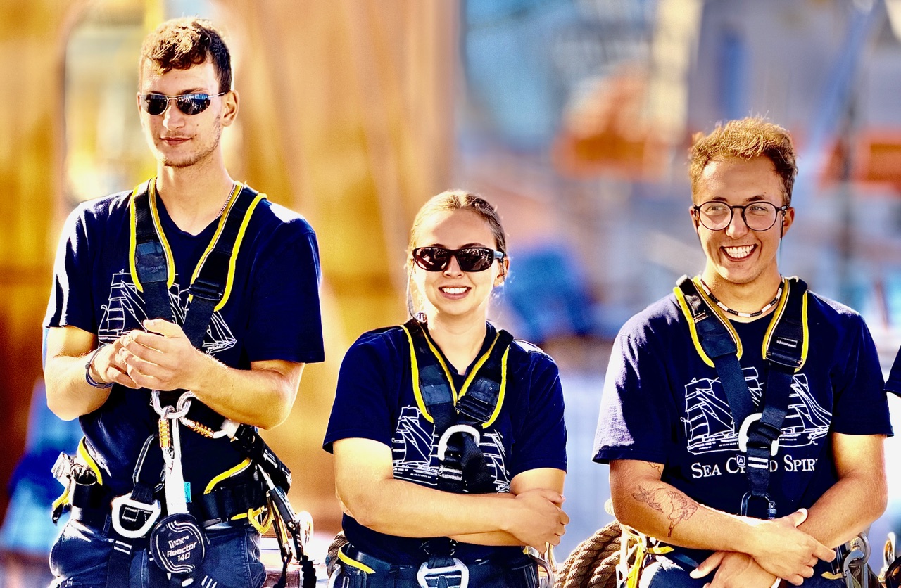 Three young crew members wearing harnesses, ready to climb the rigging.
